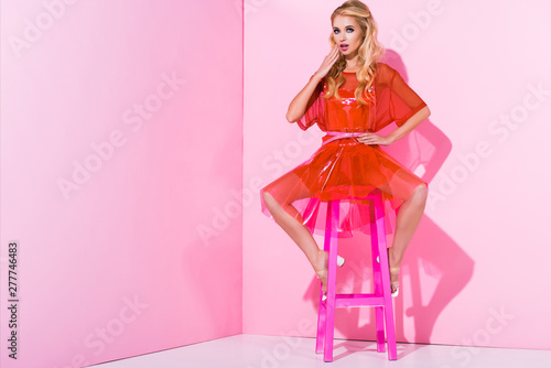beautiful girl covering mouth and posing on chair on pink, doll concept