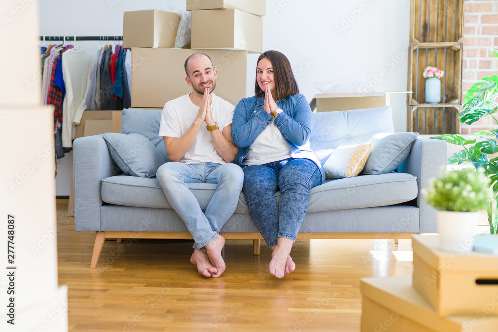 Young couple sitting on the sofa arround cardboard boxes moving to a new house praying with hands together asking for forgiveness smiling confident. Young couple sitting on the sofa arround cardboard boxes moving to a new house praying with hands together asking for forgiveness smiling confident.