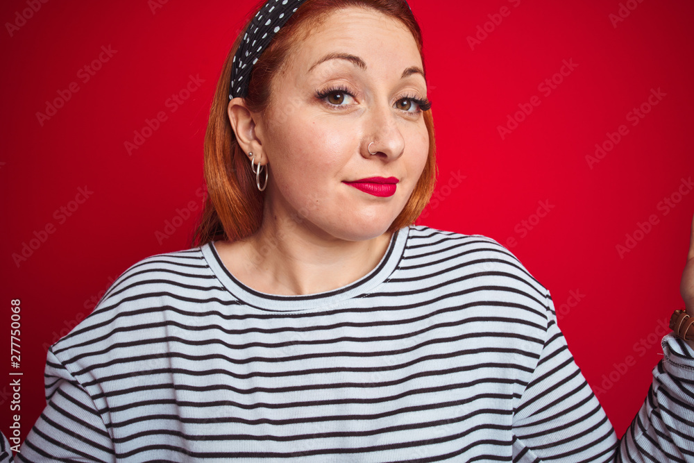 Beautiful redhead woman wearing striped navy t-shirt standing over isolated red background clueless and confused expression with arms and hands raised. Doubt concept. Beautiful redhead woman wearing striped navy t-shirt standing over isolated red background clueless and confused expression with arms and hands raised. Doubt concept.