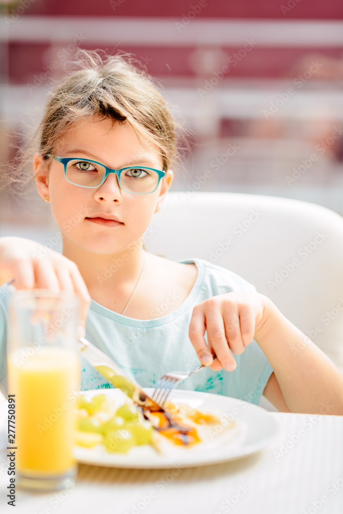 Cheerful girl eating pancakes, fresh fruits and drinking orange juice during breakfast. Healthy Lifestyle, Vegetarian Diet And Meal Cheerful girl eating pancakes, fresh fruits and drinking orange juice during breakfast. Healthy Lifestyle, Vegetarian Diet And Meal