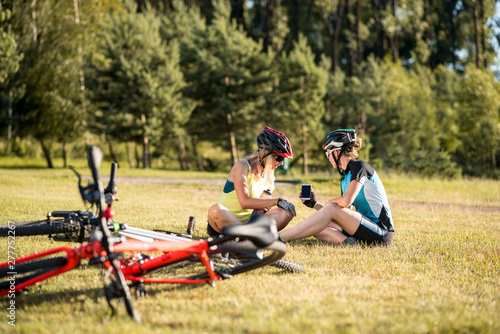 Two sports women resting on the lawn near bikes and looking to the smartphone at the map
