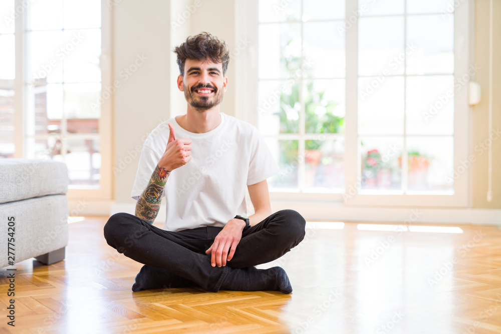 Young man working sitting casual on the floor at home doing happy thumbs up gesture with hand. Approving expression looking at the camera with showing success. Young man working sitting casual on the floor at home doing happy thumbs up gesture with hand. Approving expression looking at the camera with showing success.