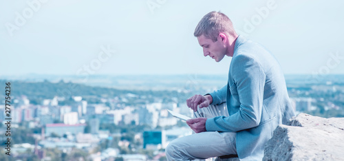 Concept: Working anytime and anywhere. Young handsome successful man is sitting on the stone and working with digital tablet over cityn from the mountains above. photo