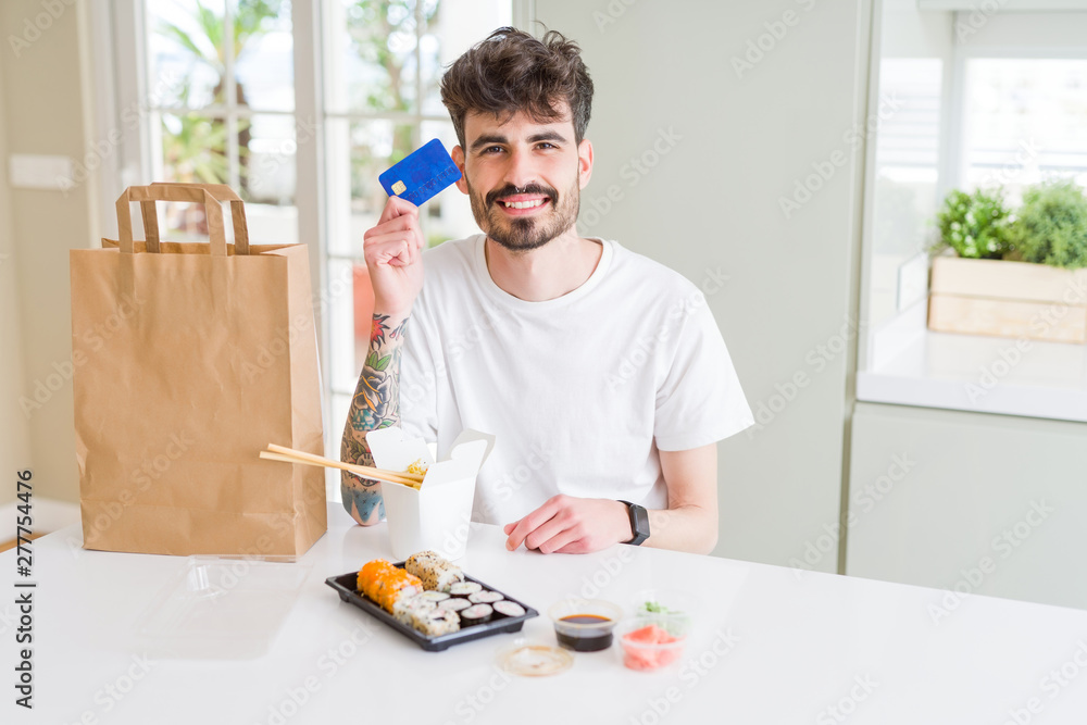 Young man eating asian sushi from home delivery using credit card as payment with a happy face standing and smiling with a confident smile showing teeth Young man eating asian sushi from home delivery using credit card as payment with a happy face standing and smiling with a confident smile showing teeth
