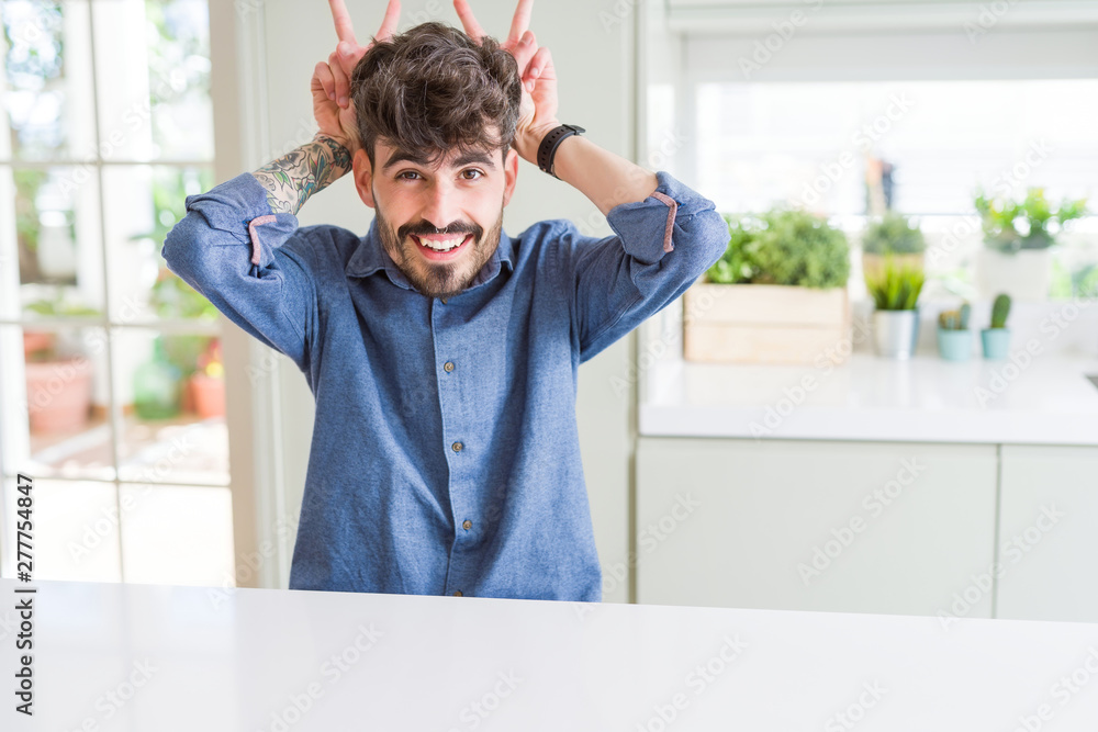 Young man wearing casual shirt sitting on white table Posing funny and crazy with fingers on head as bunny ears, smiling cheerful Young man wearing casual shirt sitting on white table Posing funny and crazy with fingers on head as bunny ears, smiling cheerful