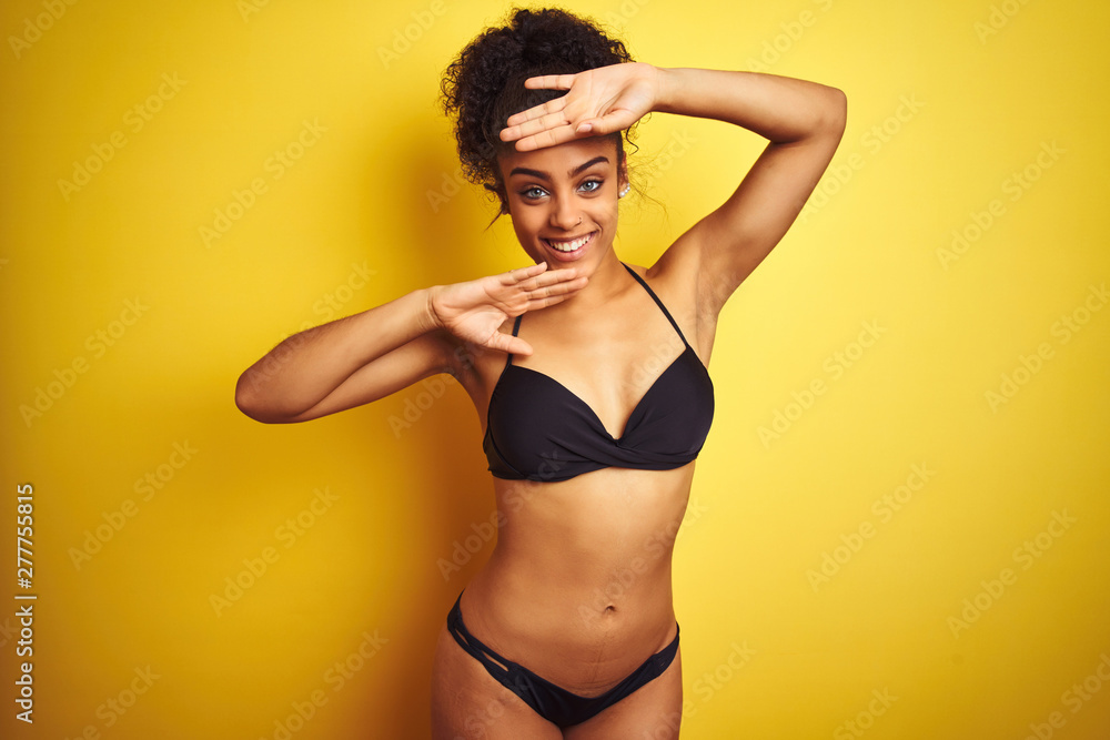 African american woman on vacation wearing bikini standing over isolated yellow background Smiling cheerful playing peek a boo with hands showing face. Surprised and exited African american woman on vacation wearing bikini standing over isolated yellow background Smiling cheerful playing peek a boo with hands showing face. Surprised and exited