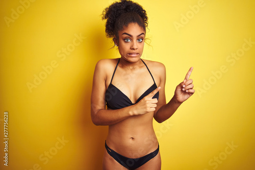 African american woman on vacation wearing bikini standing over isolated yellow background Pointing aside worried and nervous with both hands, concerned and surprised expression