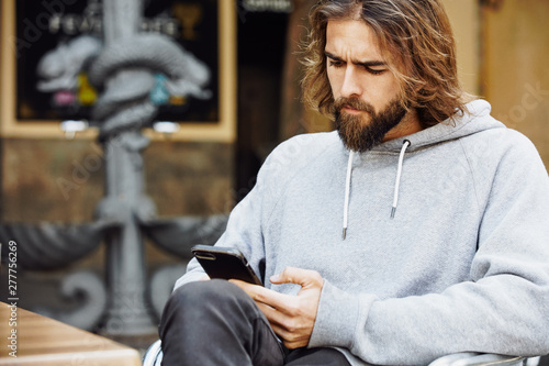 Young bearded handsome man in casual sweater sitting at cafe concentrated on mobile phone photo