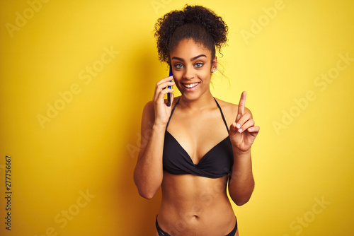 Afro american woman wearing bikini talking using smartphone over isolated yellow background surprised with an idea or question pointing finger with happy face, number one © Krakenimages.com