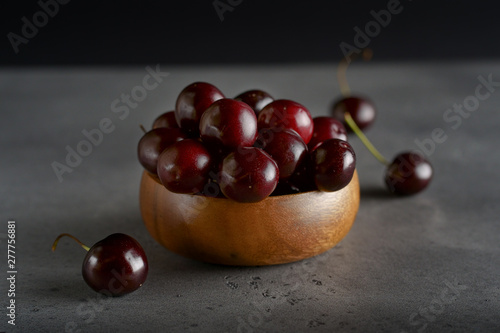 Red Cherry in a wooden cup on a gray background. some cherries are lying next to the plate. The background is dark