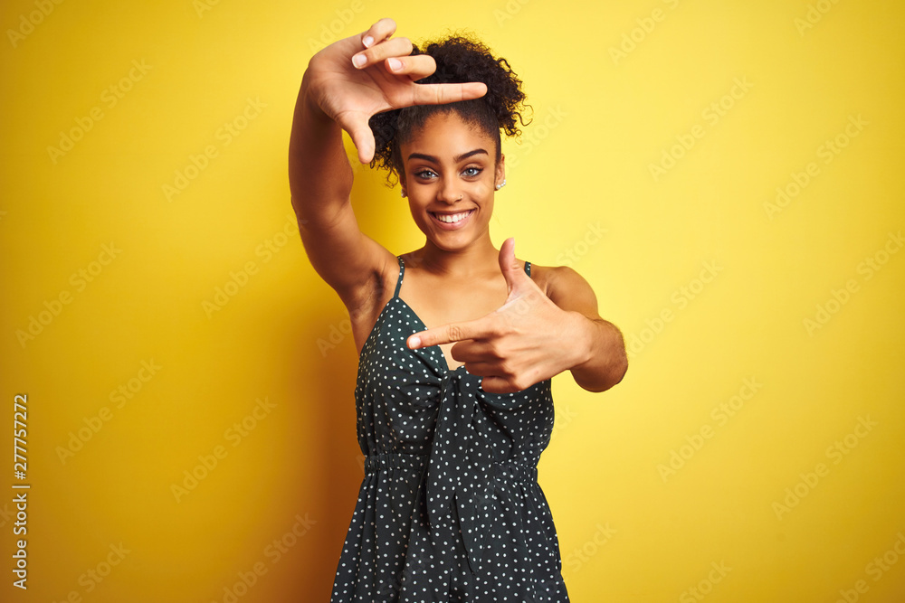 Naklejka premium African american woman wearing summer casual green dress over isolated yellow background smiling making frame with hands and fingers with happy face. Creativity and photography concept.