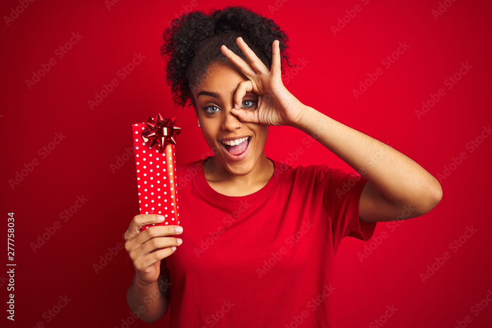 Young african american woman holding valentine gift over isolated red background with happy face smiling doing ok sign with hand on eye looking through fingers Young african american woman holding valentine gift over isolated red background with happy face smiling doing ok sign with hand on eye looking through fingers