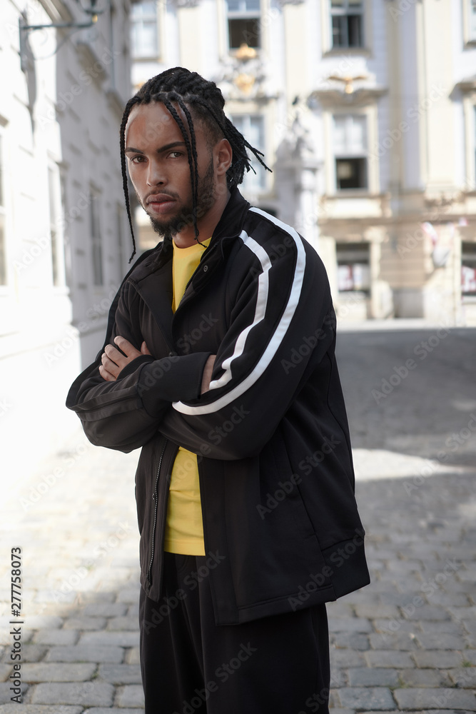 Young ethnic man with braided hair wearing black sports suit looking at camera on urban background Young ethnic man with braided hair wearing black sports suit looking at camera on urban background