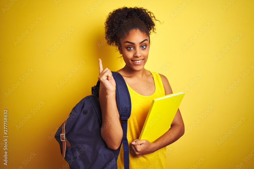 American student woman wearing backpack holding notebook over isolated yellow background surprised with an idea or question pointing finger with happy face, number one American student woman wearing backpack holding notebook over isolated yellow background surprised with an idea or question pointing finger with happy face, number one