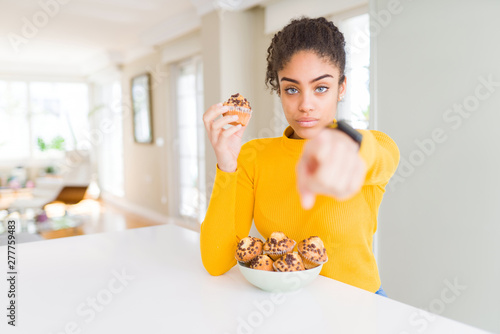 Young african american woman eating chocolate chips muffins pointing with finger to the camera and to you, hand sign, positive and confident gesture from the front