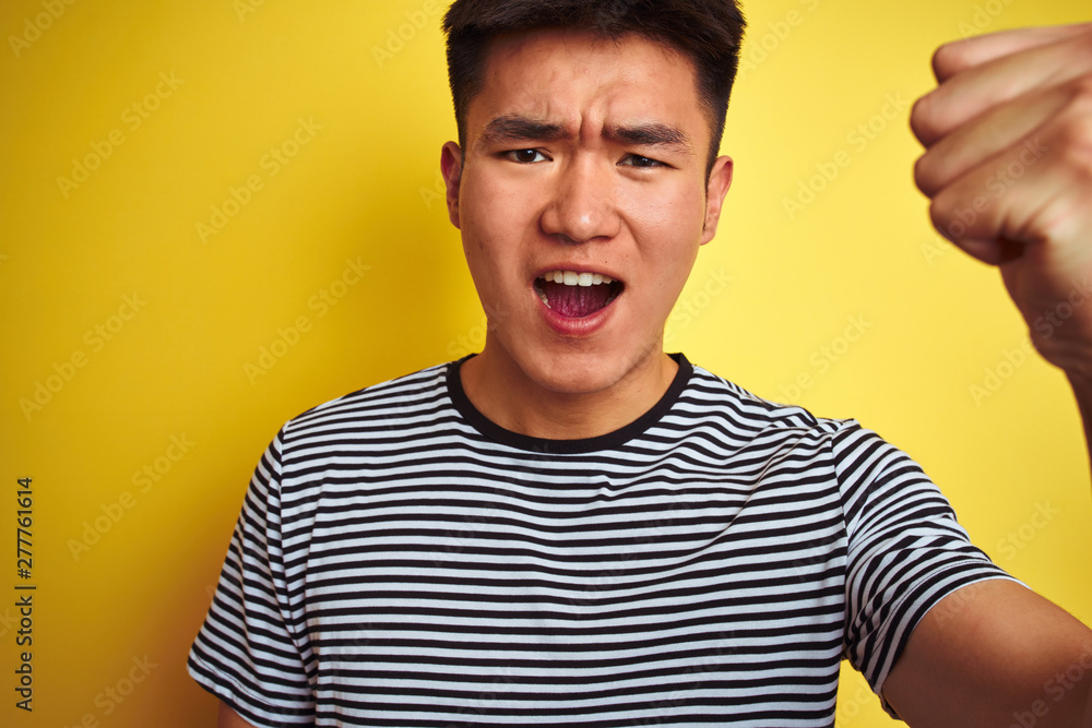 Young asian chinese man wearing striped t-shirt standing over isolated yellow background annoyed and frustrated shouting with anger, crazy and yelling with raised hand, anger concept