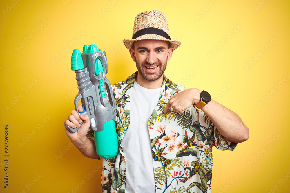 Young man wearing hawaiian flowers shirt holding water gun over yellow isolated background with surprise face pointing finger to himself Young man wearing hawaiian flowers shirt holding water gun over yellow isolated background with surprise face pointing finger to himself