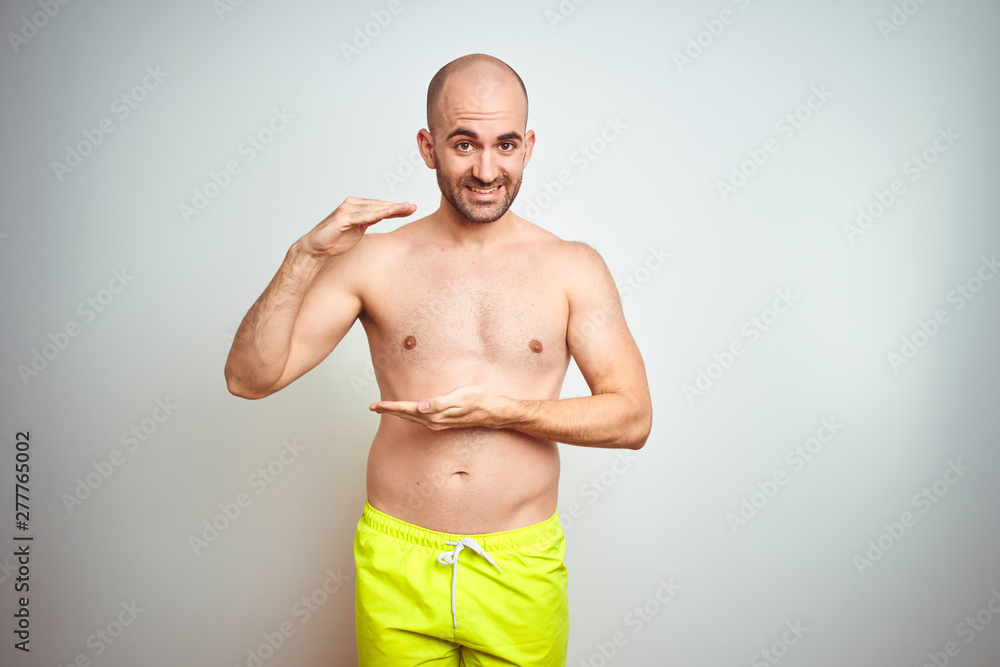 Young shirtless man on vacation wearing yellow swimwear over isolated background gesturing with hands showing big and large size sign, measure symbol. Smiling looking at the camera. Measuring concept.