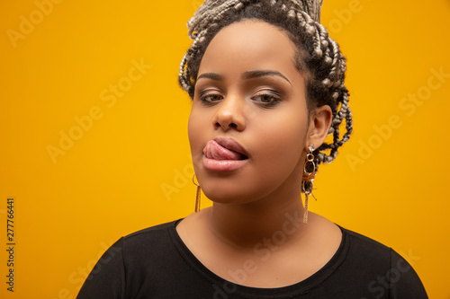 Beautiful young african american woman with dread hair showing tongue with sensuality on yellow background