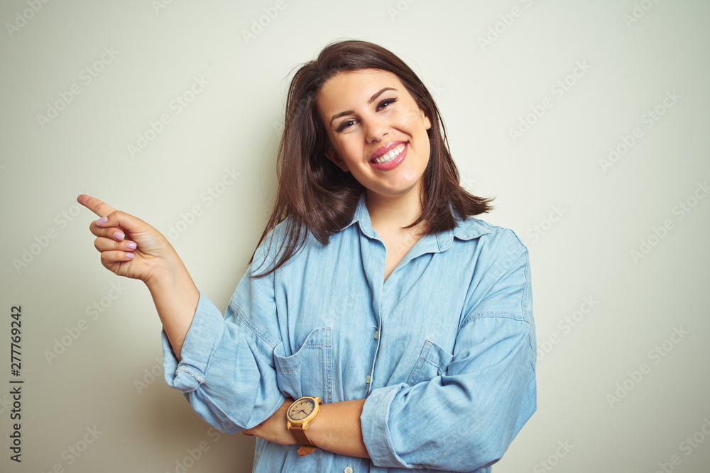 Young beautiful brunette woman wearing casual blue denim shirt over isolated background with a big smile on face, pointing with hand and finger to the side looking at the camera. Young beautiful brunette woman wearing casual blue denim shirt over isolated background with a big smile on face, pointing with hand and finger to the side looking at the camera.