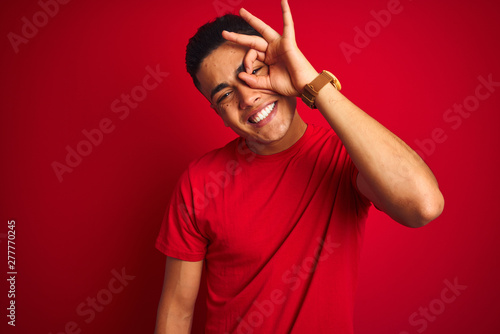 Young brazilian man wearing t-shirt standing over isolated red background doing ok gesture with hand smiling, eye looking through fingers with happy face.