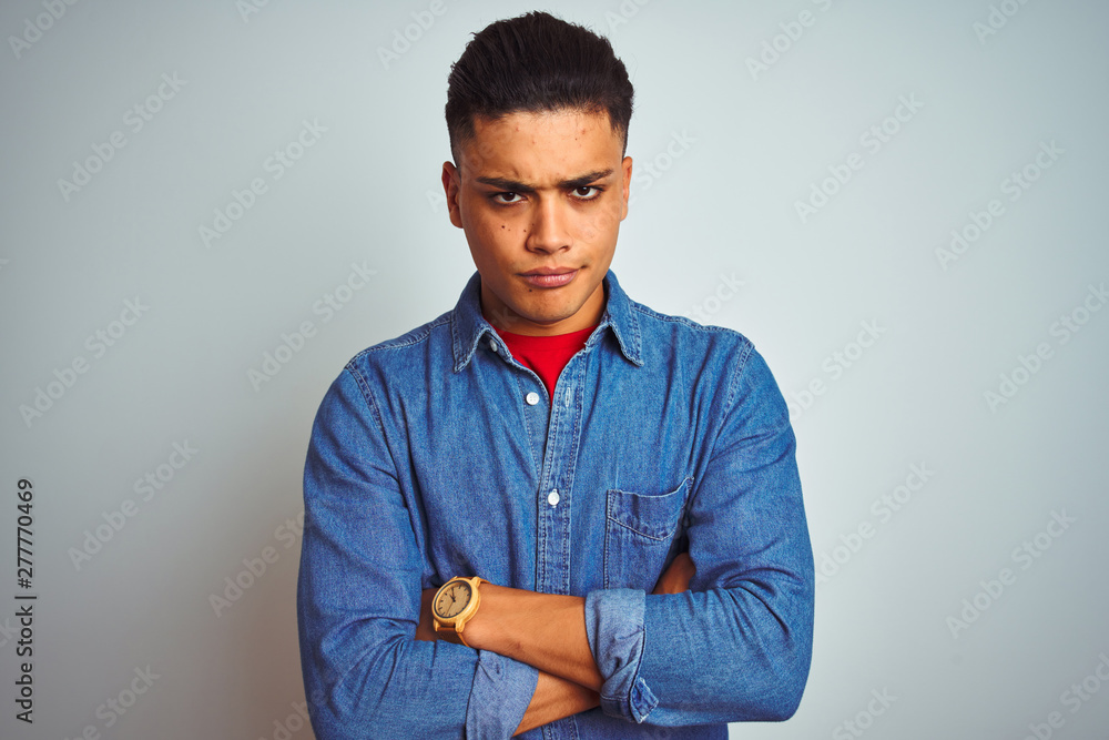 Young brazilian man wearing denim shirt standing over isolated white background skeptic and nervous, disapproving expression on face with crossed arms. Negative person. Young brazilian man wearing denim shirt standing over isolated white background skeptic and nervous, disapproving expression on face with crossed arms. Negative person.