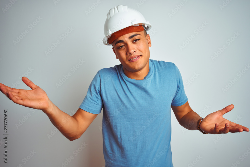 Young brazilian engineer man wearing security helmet standing over isolated white background clueless and confused expression with arms and hands raised. Doubt concept. Young brazilian engineer man wearing security helmet standing over isolated white background clueless and confused expression with arms and hands raised. Doubt concept.