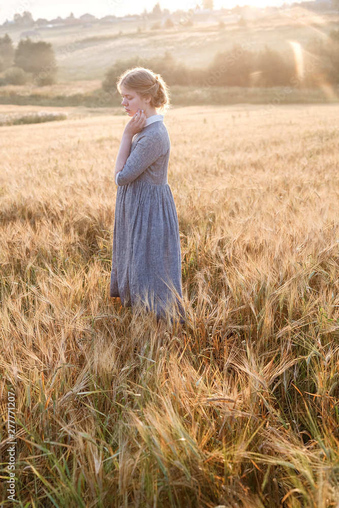 girl in blue dress standing at dawn in field of ears in golden light of sun. hill and country landscape in background girl in blue dress standing at dawn in field of ears in golden light of sun. hill and country landscape in background