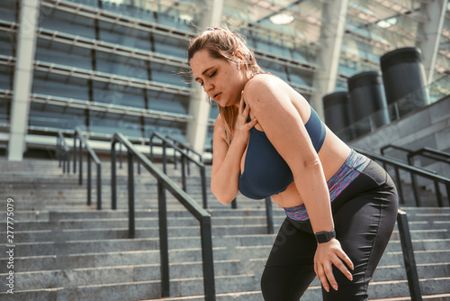 Pain after workout. Young plus size woman touching her shoulder and feeling pain after sport training while standing on stairs