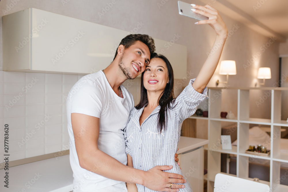 Slim dark-haired girl making selfie with husband before breakfast. Good-looking man with trendy hairstyle embracing wife which taking photo with smartphone. Slim dark-haired girl making selfie with husband before breakfast. Good-looking man with trendy hairstyle embracing wife which taking photo with smartphone.