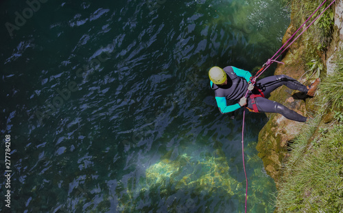 cenital view of a man wearing a neoprene sit and yellow helmet descending with strings of rappel a vertical cliff of rocks and plants to go to the water of river to canyoning in a day of summer