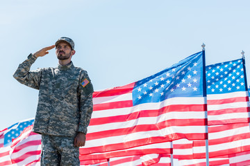 Handsome soldier in military uniform giving salute near American flags with stars and stripes