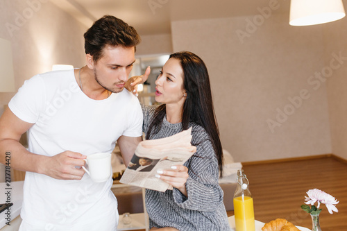 Cheerful brunette girl reading newspaper with boyfriend during lunch. Indoor portrait of serious man in white t-shirt dicussing last news with girlfriend and drinking coffee. photo