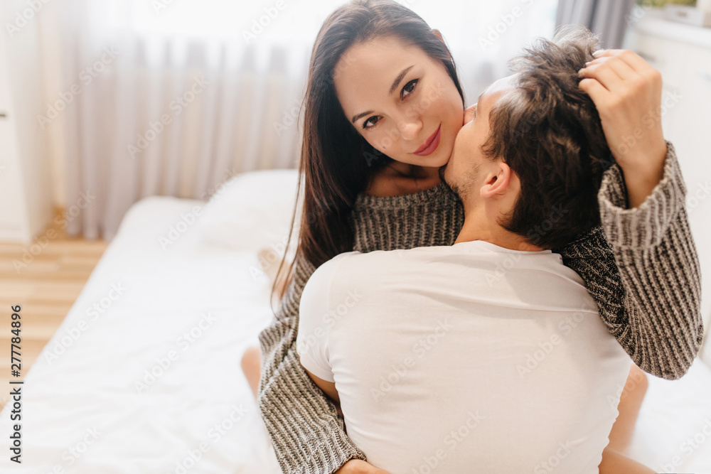 Close-up portrait from back of brunette guy embracing pretty european girl in bedroom. Indoor photo of smiling beautiful lady in woolen shirt touching husband's hair.