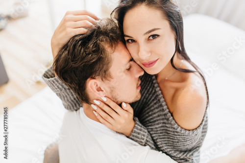 Close-up overhead photo of charming female model hugging with boyfriend in morning. Lovely girl sitting on white sheet while embracing with husband. © Look!