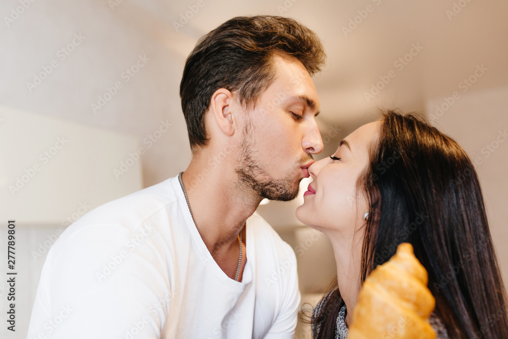 Fototapeta premium Bearded brunette man gently kissing girlfriend in nose. Indoor portrait of magnificent black-haired girl holding croissant fooling around with husband in weekend.