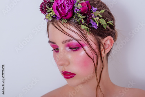 Close up beauty portrait of a girl with bright lilac professional make-up, lipstick, shadows, a hairstyle on her head and a hoop made of fresh flowers peonies, field flowers, eucalyptus. Bridesmaid