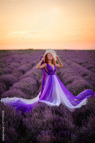 Happy red-haired woman in luxurious dress standing in lavender field at sunset