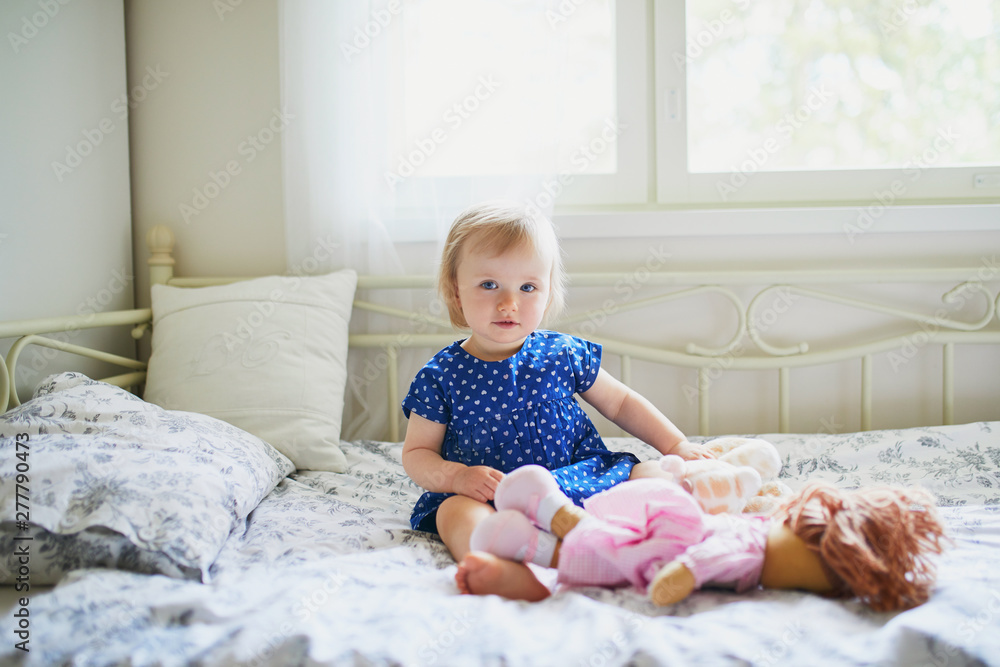 Adorable baby girl in blue dress sitting on bed and playing with a doll Adorable baby girl in blue dress sitting on bed and playing with a doll