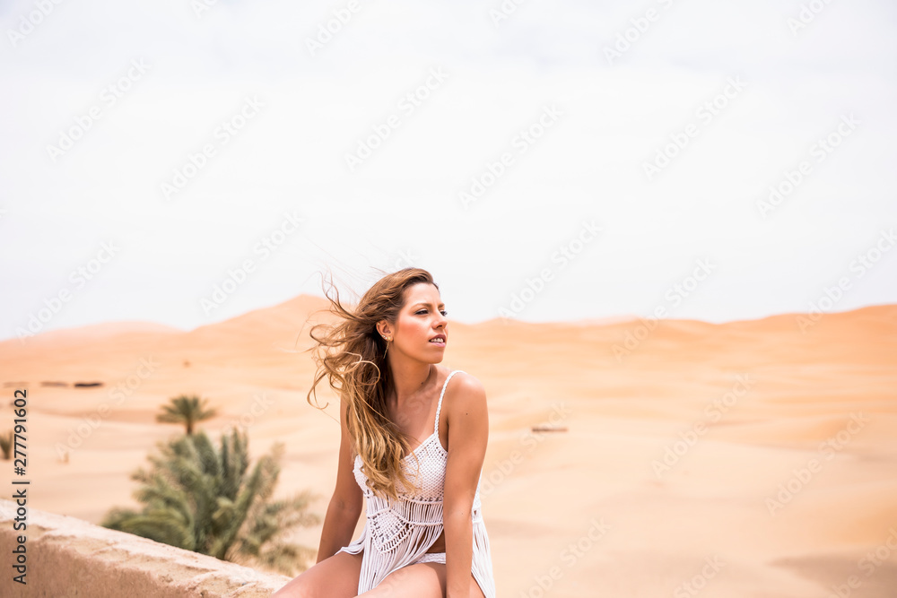 Beautiful young woman in white top sitting on stone fence in wind looking away against endless sandy desert, Morocco Beautiful young woman in white top sitting on stone fence in wind looking away against endless sandy desert, Morocco