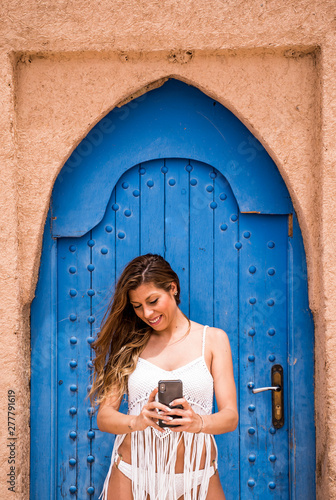 Cheerful young woman wearing white top with bikini and picturing with phone against blue oriental door in stone wall, Morocco photo