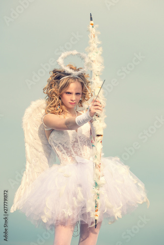 A child in the clothes of an angel on sky background. The God of Love. Valentines day. Angel wings baby. Innocent Girl with angel wings standing with bow and arrow against blue sky and white clouds
