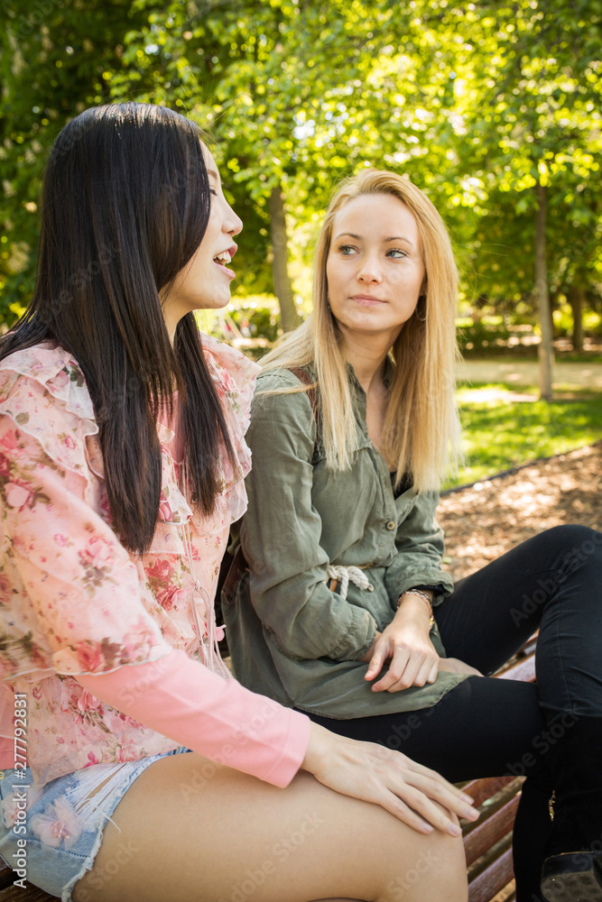 Two multiracial young women in casual outfits talking and looking at each other while sitting on bench in park Two multiracial young women in casual outfits talking and looking at each other while sitting on bench in park