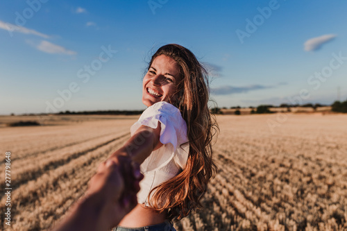 Follow me. Young woman holding hand and leading man to the beautiful nature sunset yellow landscape. View from back side, POV. Romantic couple travel, spend summer vacation together outdoors.