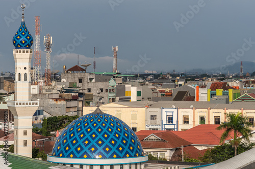 Makassar, Sulawesi, Indonesia - February 28, 2019: Masjid Babulssalam Pelabuhan mosque with dome and minaret in front of large city skyline.. Rain storn approaching cloudscape. Antennas on roofs. photo