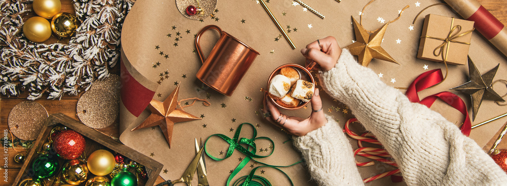 Flat-lay of woman in woolen sweater holding mug with hot chocolate drink with marshmallows, Christmas decorations, wreath, ribbons, paper, glittering balls, candy canes, top view, wide composition