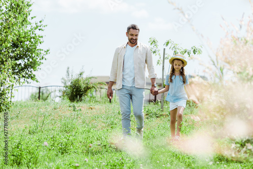 selective focus of happy father and daughter walking near green plants and holding hands