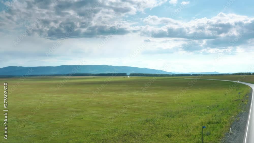 The panorama shot the terrain. Beautiful view of the field. The road crosses the field and goes through the forest towards the mountains. On the road going car. There are wonderful clouds in the sky. The panorama shot the terrain. Beautiful view of the field. The road crosses the field and goes through the forest towards the mountains. On the road going car. There are wonderful clouds in the sky.