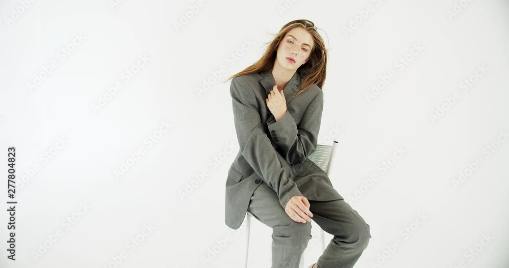 Young beautiful female model sitting on chair while posing to camera, grey stylish oversized suit Young beautiful female model sitting on chair while posing to camera, grey stylish oversized suit