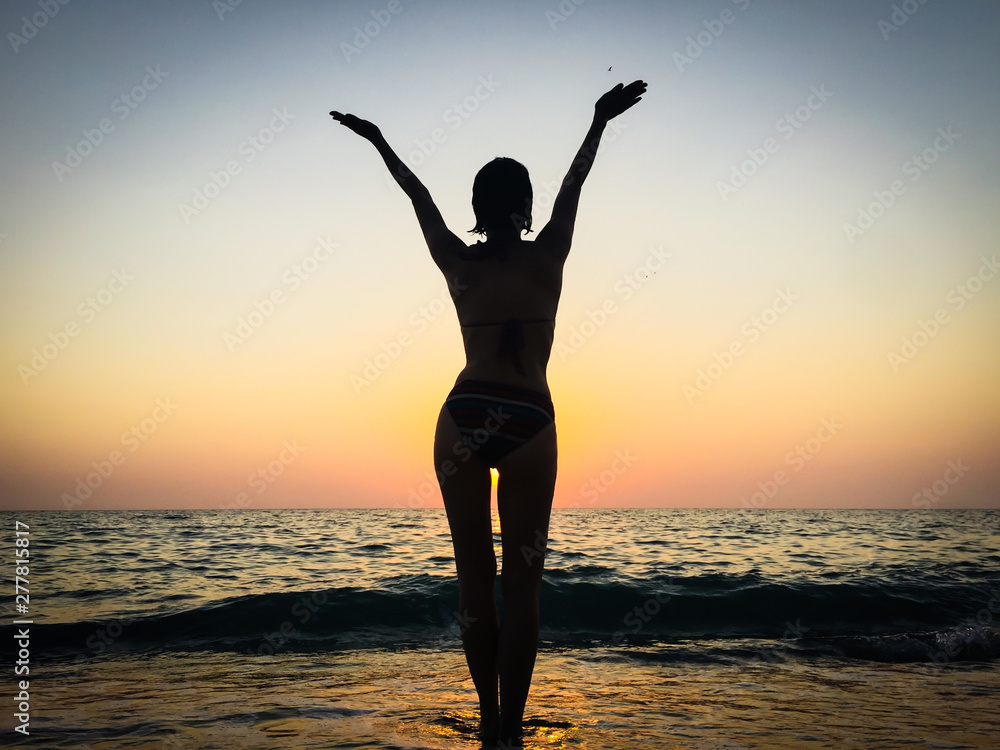 Silhouette of a slender beautiful woman against the sunset on the sea. Sexy attractive figure of a woman standing on the beach in the rays of setting sun.Girl on the beach stretches her hands up.  